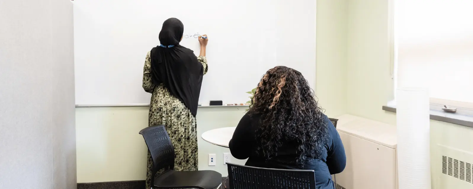 One person writes illegible text on a whiteboard while another sits and walks